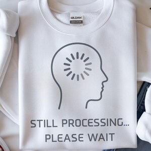 May include: White sweatshirt featuring the text "STILL PROCESSING... PLEASE WAIT" under a grey head graphic with a loading symbol. The sweatshirt is shown with blue jeans and white trainers.