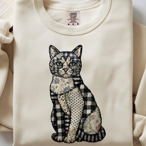 Puede incluir: Sudadera de color crema con un diseño de gato patchwork. El gato tiene un patrón de cuadros blancos y negros, detalles florales y está sentado. La sudadera tiene cuello redondo y mangas largas.