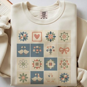 May include: Cream-colored sweatshirt featuring a patchwork design with floral, heart, and goose motifs. The design is composed of squares in muted colors, creating a vintage aesthetic. The sweatshirt has a crew neck and long sleeves.