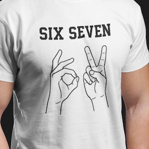 May include: White t-shirt with the text "SIX SEVEN" above hand gestures representing the numbers six and seven. The hands are drawn in black lines. The shirt is worn by a person in blue jeans.