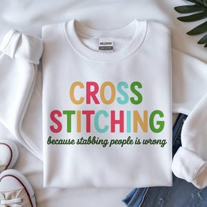 May include: White sweatshirt with the words "CROSS STITCHING" in colorful letters. Below the text is the phrase "because stabbing people is wrong" in green. The sweatshirt is paired with blue jeans and white sneakers.