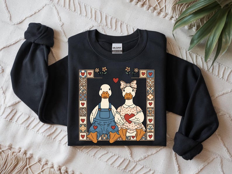 May include: Black sweatshirt featuring a charming illustration of two ducks in love. One duck wears overalls, the other a dress, both holding hearts. The design is framed with hearts and floral accents, perfect for Valentine's Day.