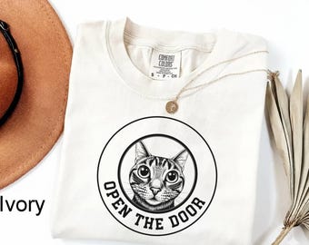 May include: Ivory-colored t-shirt with a round graphic featuring a cat's face and the text "OPEN THE DOOR" in a black circle. The shirt has a crew neck and short sleeves. A gold necklace is draped on the shirt.