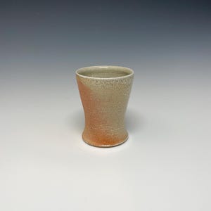 May include: A ceramic drinking glass with a light brown glaze on the bottom half and a light gray glaze on the top half.