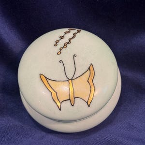 May include: A small, round, light green trinket box with a lid. The lid features a gold-outlined butterfly design with a yellow center. Above the butterfly are gold-colored, chain-like accents. The box is likely ceramic or porcelain.