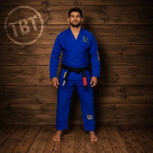 May include: A royal blue Brazilian Jiu-Jitsu gi featuring a long-sleeve jacket with a V-neck collar and matching trousers. The uniform includes a black belt with a red tab and gold embroidered logos on the chest, sleeve, and pant leg.
