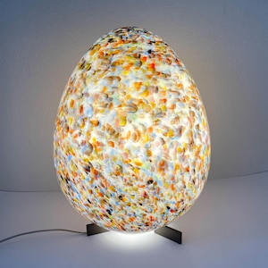 Peut inclure: Lampe de table en forme d'œuf avec un motif tacheté et coloré. La lampe a un socle à finition sombre et un cordon. L'abat-jour est un mélange de jaune, d'orange, de bleu et de blanc, créant un aspect vibrant et artistique. Hauteur environ 30 cm.