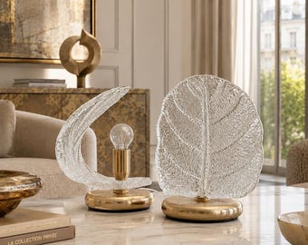 May include: Two table lamps featuring textured clear glass shades shaped like leaves, one curved and one upright, mounted on circular gold-toned metal bases. The lamps include exposed light bulb sockets and gold-toned hardware, creating a reflective look.