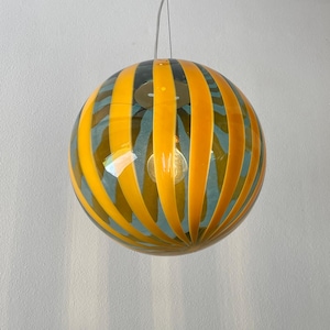 Op de afbeelding: Een bolvormige glazen hanglamp met afwisselende verticale strepen van ondoorzichtig geel en doorschijnend blauw glas. De bol hangt aan een dunne metalen draad en bevat een enkele zichtbare gloeilamp in het midden van de armatuur.