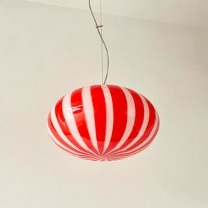 May include: A red and white striped pendant light fixture. The light has an oval shape with vertical stripes. The fixture is suspended from the ceiling by a thin wire and a gold-colored canopy.