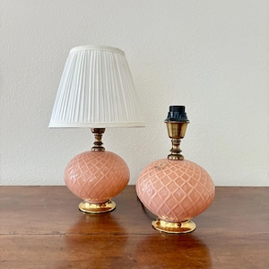 May include: Two matching table lamps. One lamp has a white pleated lampshade. Both lamps have a peach-colored, textured, spherical base with a gold-colored base and brass-colored accents. The lamps are on a wooden surface.