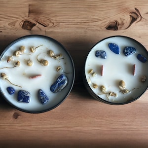 May include: Two white candles with blue and red stones embedded in the wax. The candles are in black containers and are on a wooden surface.