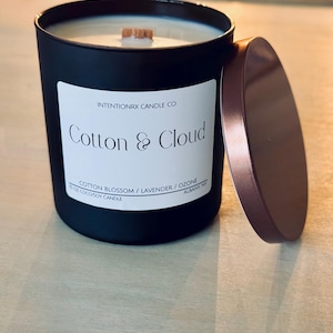 May include: A black candle jar with a wooden lid. The label reads "INTENTIONRX CANDLE CO. Cotton & Cloud COTTON BLOSSOM/LAVENDER/OZONE 10 OZ COCOSOY CANDLE ALBANY NY"