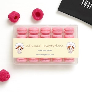 May include: A clear plastic box filled with pink macarons, arranged in rows. The box has a cream-colored label with the text "Almond Temptations" and a cartoon chef illustration. Fresh raspberries are scattered around the box. The website address almondtemptations.com is also visible.