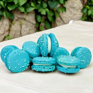 May include: A collection of light blue macarons with white filling and white sprinkles. Some macarons are open, revealing the creamy filling. The macarons are arranged on a white surface, with green foliage in the background.