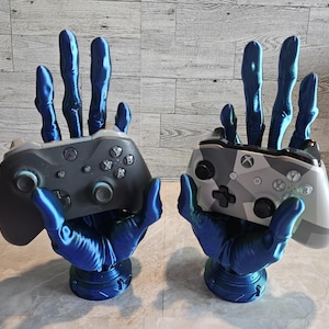 May include: Two blue 3D printed hands holding video game controllers. The controllers are black and white with buttons and a directional pad. The hands are posed as if they are holding the controllers in a playing position.