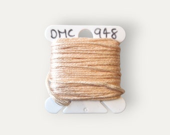 Puede incluir: Una bobina de hilo de bordar DMC en color 948, un beige claro. El hilo está enrollado en una bobina de plástico blanca con el logotipo de DMC y el número de color impreso en ella.