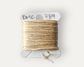 Puede incluir: Una bobina de hilo de bordar DMC en color 739, un color beige claro. La bobina está etiquetada con el logotipo de DMC y el número de color.