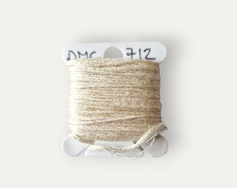 Puede incluir: Una bobina de hilo de bordar DMC de color beige claro, etiquetada con el número "712".