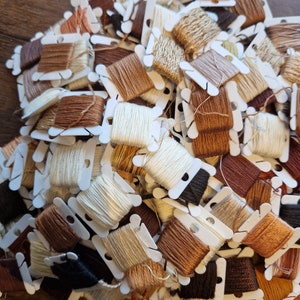 May include: A pile of small spools of embroidery floss in various shades of brown, beige, and white. The spools are arranged in a random pattern and are all in plastic containers.