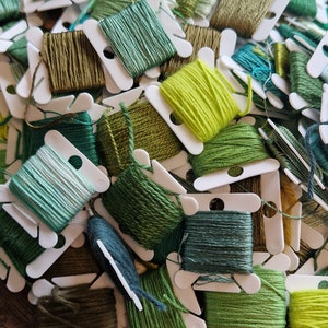 Può includere: Una collezione di rocchetti di filo da ricamo verde, blu e giallo in bobine di plastica bianca. I rocchetti sono disposti in una pila, mostrando una varietà di tonalità di verde.