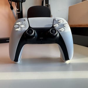 May include: A white and black PlayStation 5 DualSense wireless controller. The controller has a white faceplate with black accents, two analog sticks, and various buttons. The controller is designed for gaming.
