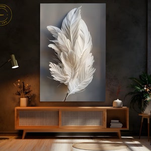 Könnte beinhalten: Ein minimalistischer Wandkunstdruck mit einem großen weißen Federdesign auf grauem Hintergrund. Der Druck ist gerahmt und hängt an einer Wand über einem Holzschränk.