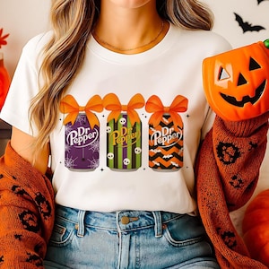 May include: White t-shirt featuring three Halloween-themed Dr. Pepper cans with orange bows. The cans have different designs: purple with spiderwebs, green with skulls, and orange and black zigzags. A pumpkin-shaped container is held in the background.