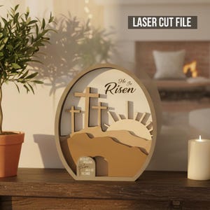 Op de afbeelding: Een lasergesneden houten paasdecoratie met drie kruisen op een heuvel, met de tekst "He Is Risen". Het ontwerp bevat een opkomende zon en een graf. De lijst is ovaal van vorm.