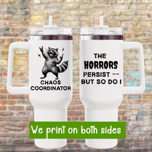 May include: White travel mug with a handle, featuring a raccoon graphic with the text "CHAOS COORDINATOR" on one side and "THE HORRORS PERSIST -- BUT SO DO I" on the other. The mug has a clear lid and a green banner that says "We print on both sides".