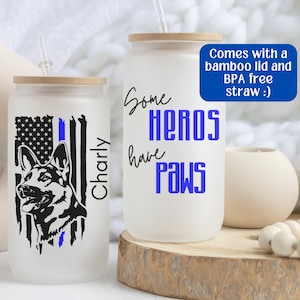 May include: Two white tumblers with a bamboo lid and a blue straw. One tumbler has a black and white silhouette of a German Shepherd with an American flag and a blue line. The other tumbler has the text "Some Heros Have Paws".