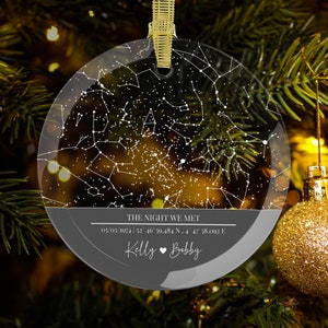 May include: A clear glass ornament with a black and white star map design. The ornament has the text "THE NIGHT WE MET" and coordinates "05.05.1074 | 52 46' 39.484 N, 4 47' 38.092 E". The ornament also has the names "Kelly" and "Bobby" with a heart symbol between them.
