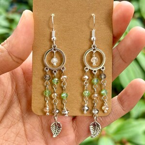 May include: Silver dangle earrings with a circle charm and a leaf charm. The earrings are decorated with green, clear, and brown beads.