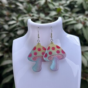 May include: A pair of mushroom-shaped earrings. The earrings feature a light yellow cap with red polka dots, a pink underside, and a blue-green stem. The earrings are displayed on a white mannequin head against a blurred green background.