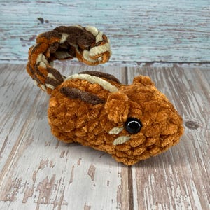 May include: A handmade, plush chipmunk toy in shades of brown, orange, and cream. The chipmunk has a striped back, a curled tail, and a black button eye. The toy is made of soft, textured yarn.