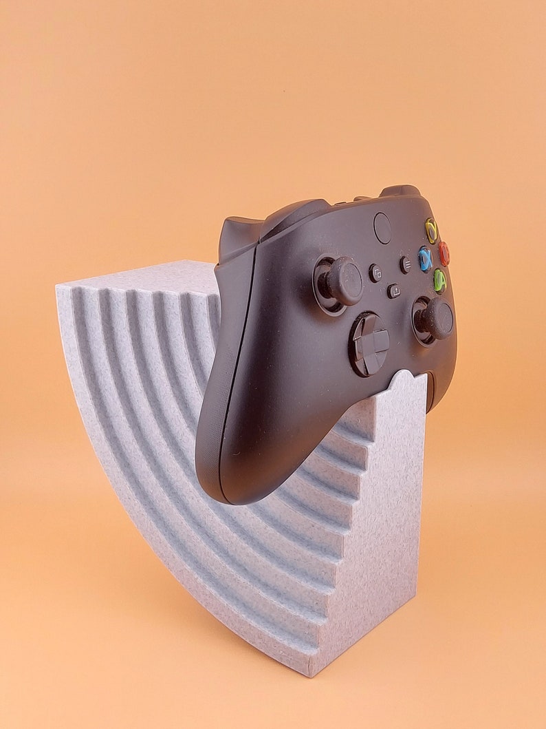 以下が含まれることがあります： 黒いビデオゲームコントローラーが、グレーのテクスチャ加工された湾曲したスタンドの上に置かれています。コントローラーは黒と白のデザインで、緑、青、赤のボタンが付いています。