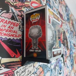 May include: A silver Pop Rocks collectible figure of Corey Taylor, the lead singer of Slipknot, wearing a black jacket and black trousers. The figure is in a clear plastic box with a red and black background. The box features the Pop Rocks logo and the number 326.