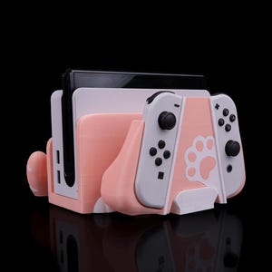 May include: A pink and white Nintendo Switch console stand with a paw print design. The stand holds the console and two controllers.