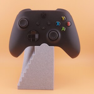 以下が含まれることがあります： グレーのスタンドが付いた黒いXboxコントローラー。コントローラーには、白黒の十字キー、白黒のアナログスティック、そして色付きの文字（X、Y、A、B）が書かれた黒いボタンがあります。