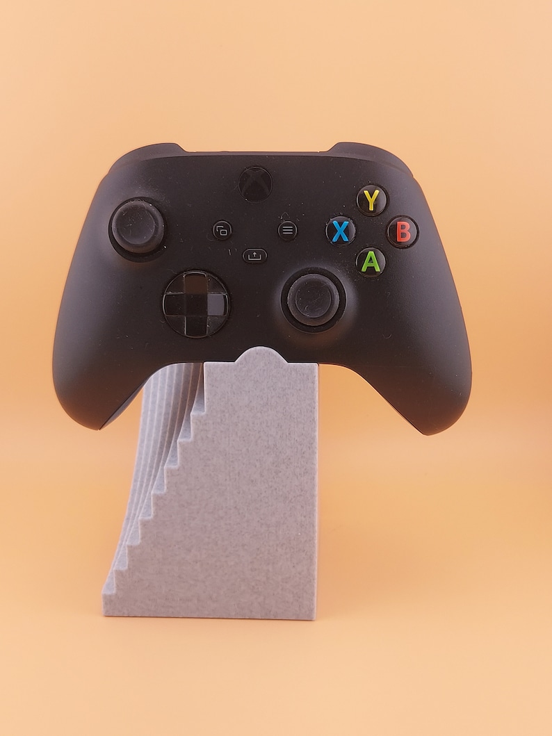 以下が含まれることがあります： グレーのスタンドが付いた黒いXboxコントローラー。コントローラーには、白黒の十字キー、白黒のアナログスティック、そして色付きの文字（X、Y、A、B）が書かれた黒いボタンがあります。