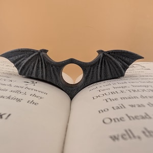 May include: A black bat-shaped bookmark with a hole in the centre, resting on an open book. The bookmark is made of a textured material and has a slightly worn look.
