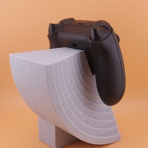 以下が含まれることがあります： 黒いビデオゲームコントローラーが置かれた、グレーの湾曲した多層スタンド