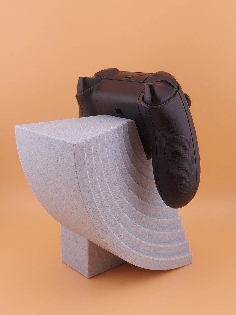 以下が含まれることがあります： 黒いビデオゲームコントローラーが置かれた、グレーの湾曲した多層スタンド