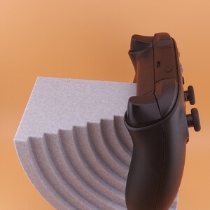 以下が含まれることがあります： グレーのボタンが付いた黒いビデオゲームコントローラーが、グレーのテクスチャのある湾曲した表面の上に置かれています