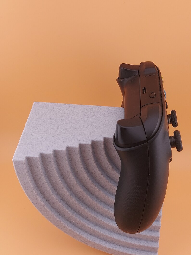 以下が含まれることがあります： グレーのボタンが付いた黒いビデオゲームコントローラーが、グレーのテクスチャのある湾曲した表面の上に置かれています