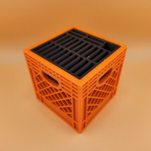 May include: An orange and black miniature crate with a slotted top. The crate has a square shape with rounded cutouts on the sides. The top is black with parallel slots.