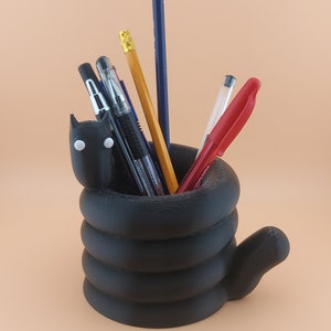 Może przedstawiać: Czarny organizer na biurko w kształcie kota ze spiralnym ciałem i ogonem. Organizer jest wypełniony długopisami i ołówkami, w tym czerwonym długopisem, niebieskim długopisem, czarnym długopisem i żółtym ołówkiem.