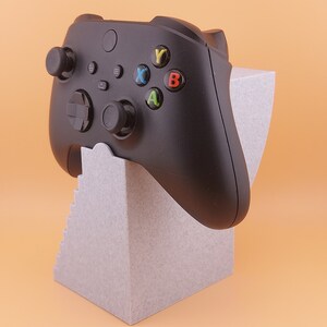 以下が含まれることがあります： 黒いビデオゲームコントローラーに色付きのボタンが付き、灰色のスタンドに置かれています。スタンドはテクスチャ加工が施され、段状のデザインになっています。