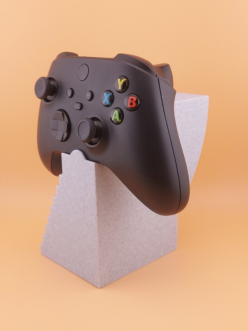 以下が含まれることがあります： 黒いビデオゲームコントローラーに色付きのボタンが付き、灰色のスタンドに置かれています。スタンドはテクスチャ加工が施され、段状のデザインになっています。
