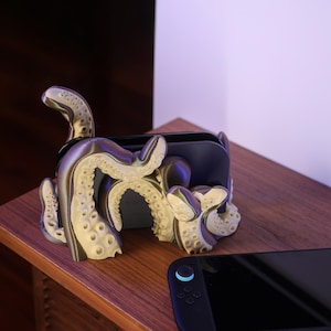 Op de afbeelding: Een unieke, tentakelachtige telefoon- of gamecontrollerhouder in crème en grijs. De houder is ontworpen als een octopus en staat op een houten oppervlak. Een zwarte gamecontroller ligt naast de houder.